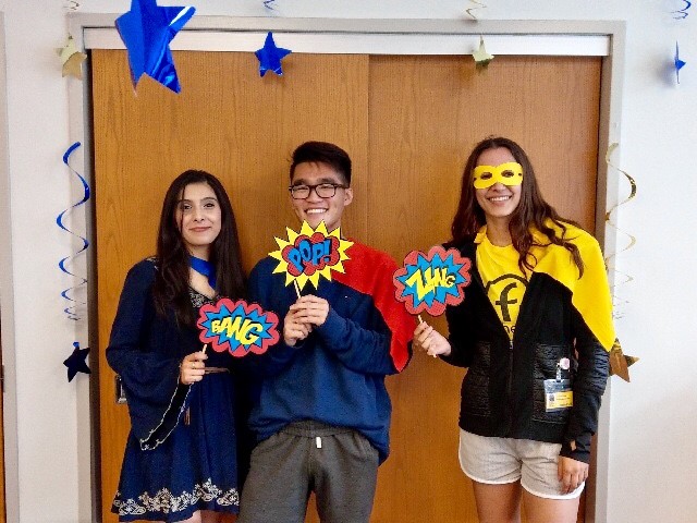 Three students stand next to each other. They hold colorful signs that say 'pop!' 'boom!' and 'zap!'